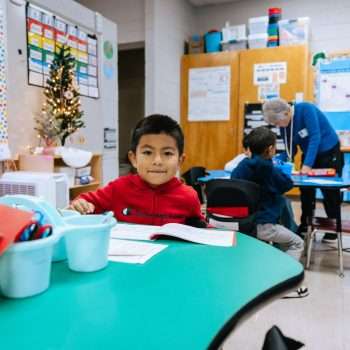 Little boy wearing a red sweatshirt is sitting behind a green table holding a pencil.